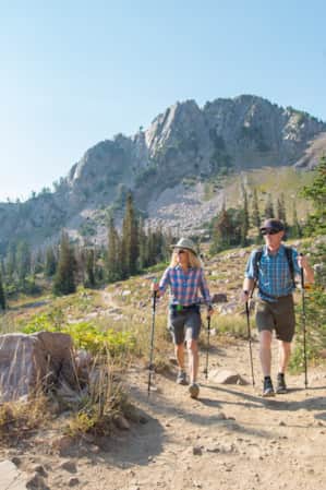 A man and woman hiking with poles on a dirt trail, with a large mountain peak behind them.