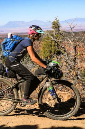 A woman biking through the desert on a sandy trail, with greenery on one side.
