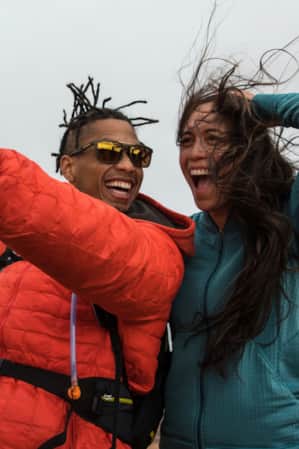 Two people hiking in the desert, smiling at their phone camera for a selfie while the wind blows their hair around.