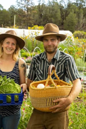 A man and a woman each holding a basket of vegetables, with a garden visible behind them.