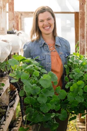 A woman smiling while holding small crates of plant sprouts inside a greenhouse.