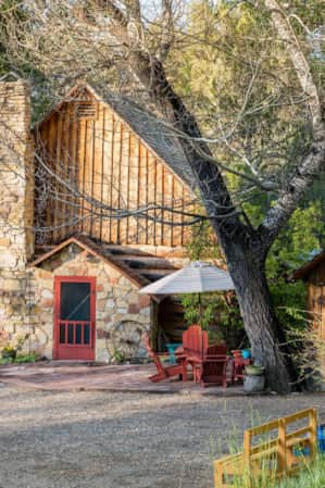 A rustic log cabin with a stone chimney, a red door, and a small wooden shed, surrounded by trees with a seating area featuring red Adirondack chairs under a white umbrella in front.