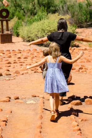 A woman and two children walk together with arms outstretched along a winding path made of stones in a desert garden, surrounded by greenery.