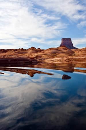 A lake reflecting a blue sky with a few clouds, surrounded by hills of red sandstone.
