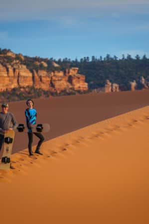Two people are smiling and holding sandboards to surf down a sand dune at Coral Pink Sand Dunes State Park.