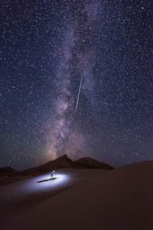 A man standing on a sand hill with a flashlight, looking up at a dark sky filled with stars and the Milky Way.