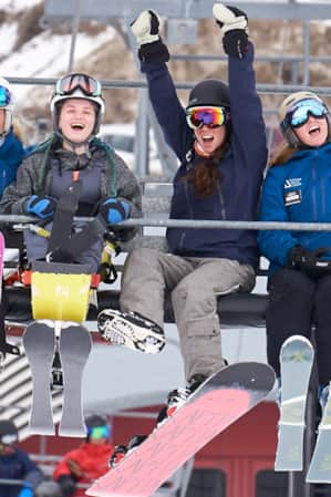 Four women on a ski lift, dressed in winter clothes and ski gear, are smiling and excitedly throwing their hands up in the air.
