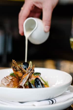A bowl of elegantly displayed seafood, being topped with a white sauce.