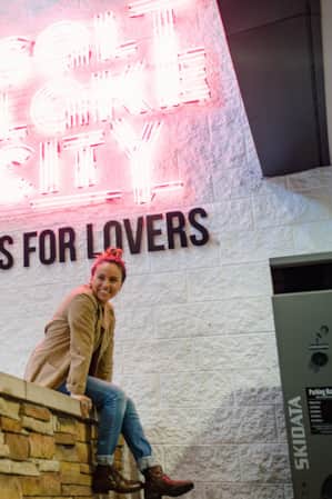 A woman sitting and smiling at the base of a sign that reads 'Salt Lake City is for lovers,' while having her picture taken.