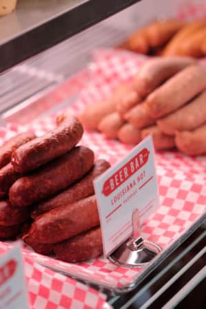 A deli case displaying various types of sausage links on silver trays lined with red and white checkered paper.