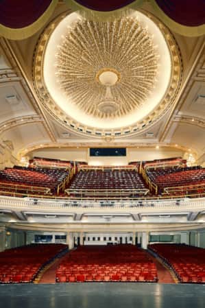 The view from the stage of a theater, showcasing ornately decorated ceilings and red theater chairs.