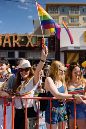 A crowd of people outside celebrating Pride with flags and rainbow clothing in front of two bars.