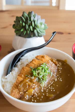 A bowl of rice topped with thick brown curry sauce, placed on a wooden table in a restaurant.