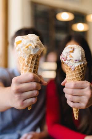 Two people out of focus, each holding up ice cream cones that are in focus, covering their faces.