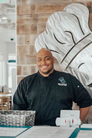 A man wearing a chef's uniform, smiling behind the counter at a restaurant.