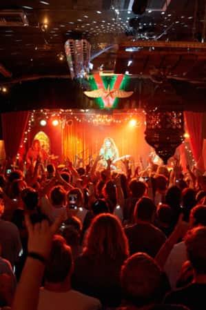 A crowded room with red lights, where people are looking at and listening to a live band in an ornate setting.