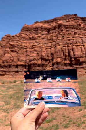A person holding a photograph in front of a red rock formation, with the photograph depicting a scene from a movie shot at the same location where the person is standing.