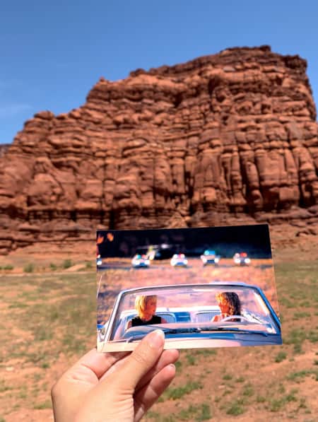 A person holding a photograph in front of a red rock formation, with the photograph depicting a scene from a movie shot at the same location where the person is standing.
