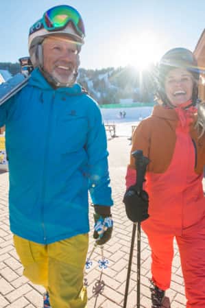 A man and a woman smiling in ski gear, carrying their skis on their shoulders through a ski resort.