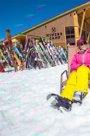 A man and a woman sitting outside on lawn chairs at a mountain lodge, both wearing ski gear.