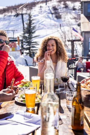 People at a ski resort, bundled up in winter clothing, eating and drinking at a wooden table outside.