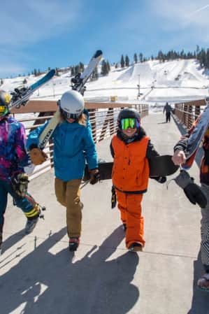 Four kids walking side by side, carrying skis and snowboards as they cross a bridge, with ski slopes in the background.
