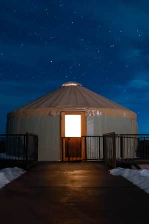A yurt surrounded by a small fence, situated in the middle of a snowy landscape under a starry night sky.